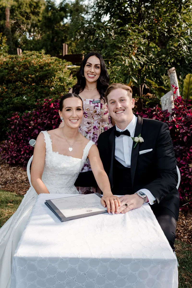 Ellie smiling behind a couple signing their marriage certificate in a garden